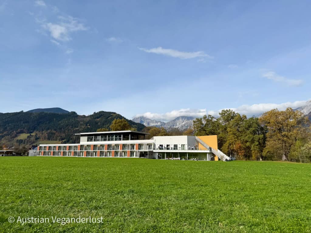 Ein modernes Hotelgebäude mit klarer, kantiger Architektur vor der malerischen Kulisse der österreichischen Alpen. Die üppige grüne Wiese im Vordergrund verleiht dem Ort eine heitere Note.
