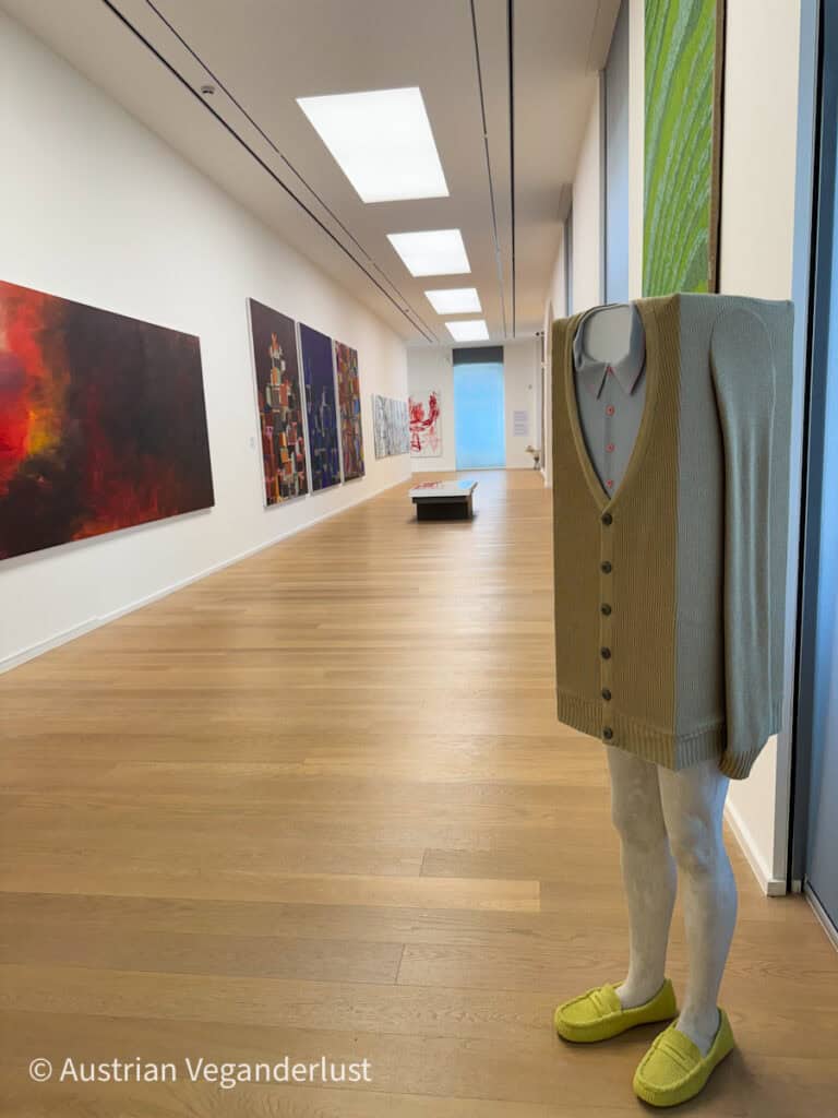 A contemporary art exhibit at Admont Modern Museum featuring abstract paintings on white walls and a unique sculpture of a headless figure wearing a sweater and slippers, creating a minimalist and avant-garde atmosphere.