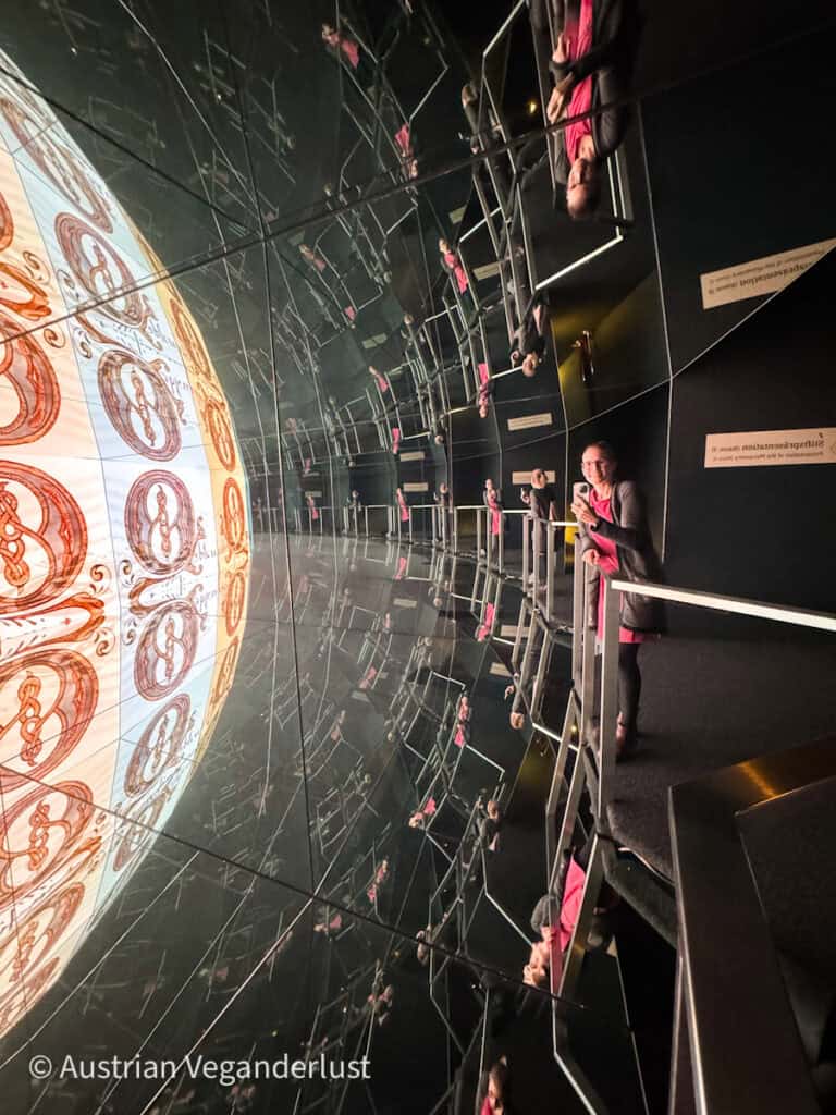 A visitor interacting with a mirrored exhibit at Admont Multimedia Museum, where a large, reflective wall displays intricate designs and the visitor’s reflection creates an immersive experience.