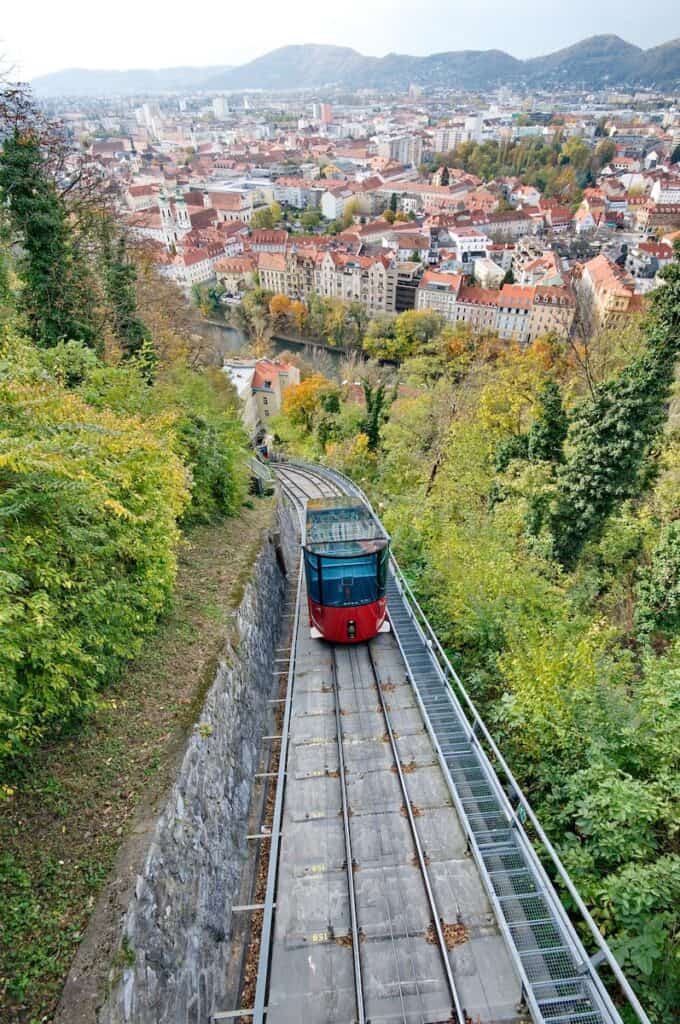 A red funicular car ascends the steep Schlossbergbahn railway in Graz, offering a scenic view of the city’s rooftops and surrounding mountains. The ride is a popular way to reach the top of the Schlossberg hill.