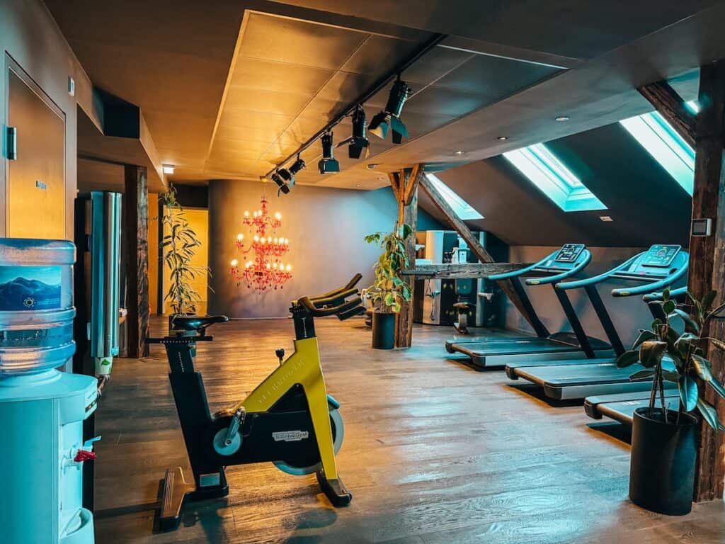 Ein Hotel-Fitnessraum mit einem gelben Heimtrainer, Laufbändern und einem Wasserspender. Der Raum hat Holzbalken, schräge Decken mit Oberlichtern und dekorative Pflanzen.