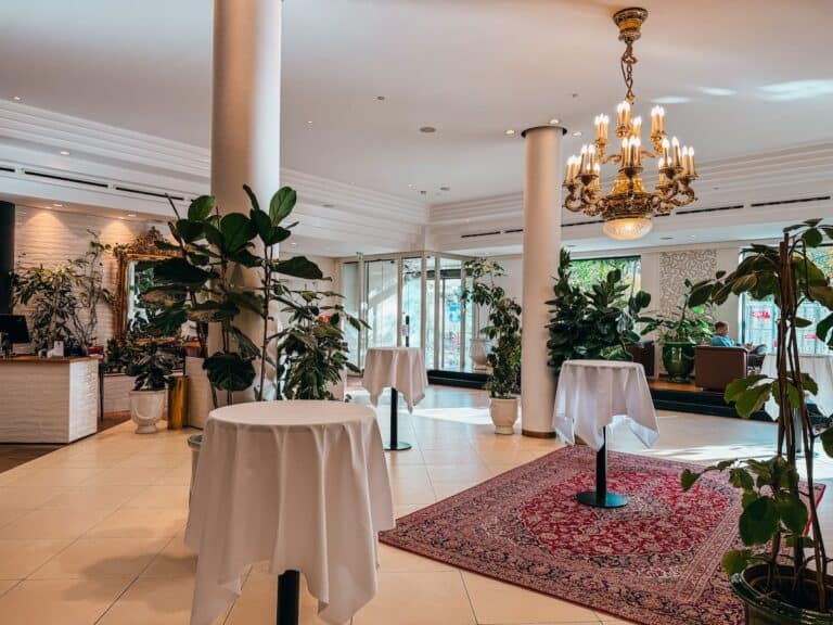 Die Lobby des Weitzer Hotels mit runden, mit weißen Tüchern bedeckten Cocktailtischen, einem großen Kronleuchter und zahlreichen Topfpflanzen. Der Raum ist hell und ein roter Teppich verleiht ihm einen Hauch von Eleganz.