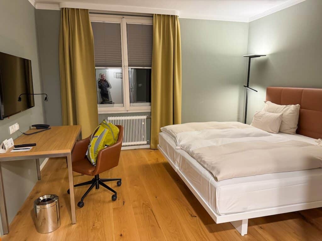 Ein Hotelzimmer mit einem großen Doppelbett, Holzboden und einem Schreibtisch mit Lederstuhl. Das Zimmer hat eine sanfte Beleuchtung, neutrale Vorhänge und ein modernes, minimalistisches Design.