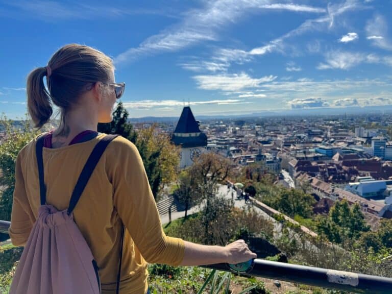 Tina mit Rucksack bewundert den Panoramablick auf Graz vom Schlossberg aus. Im Hintergrund ist der Uhrturm zu sehen, umrahmt von einem klaren Himmel und einem weiten Stadtbild.