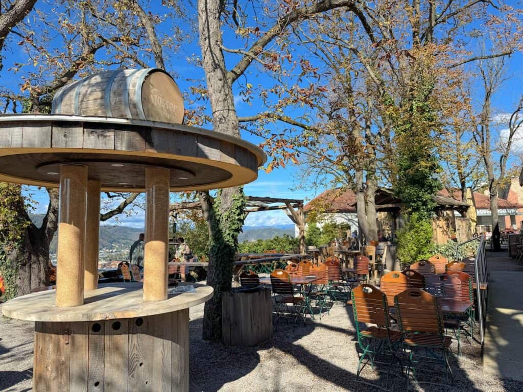 A traditional outdoor beer garden on Schlossberg hill, featuring wooden picnic tables and barrel-shaped decorations under the shade of trees. The relaxed atmosphere invites visitors to enjoy local beverages.