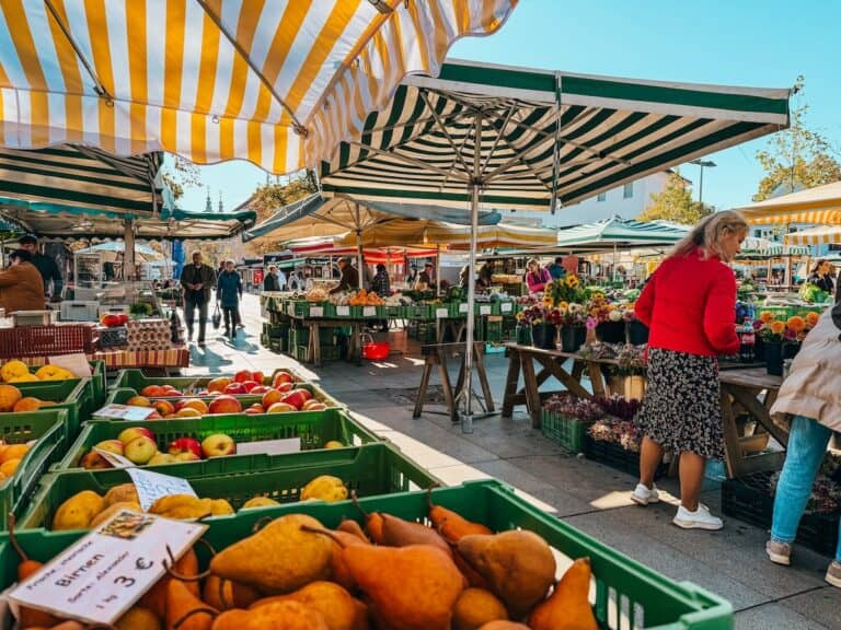 A bustling outdoor market in Graz, Austria, with colorful striped canopies sheltering stalls filled with fresh produce, flowers, and handmade goods. Shoppers explore the vibrant selection of fruits, vegetables, and artisanal products.