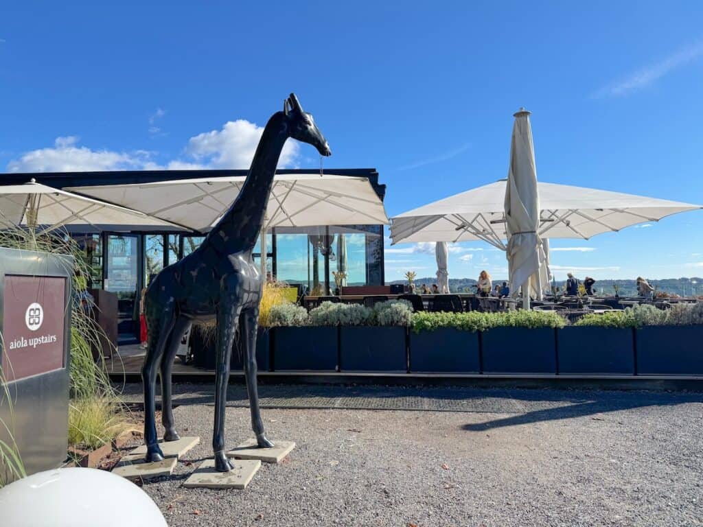 An outdoor seating area of the Aiola Upstairs Café on Schlossberg hill in Graz, with large umbrellas and a modern, sleek design. A statue of a giraffe stands near the entrance, adding a whimsical touch.
