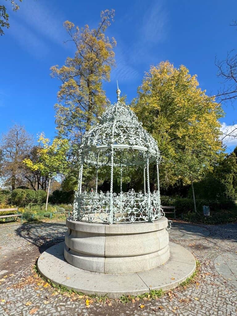 A decorative wrought-iron fountain on Schlossberg hill in Graz, featuring intricate metalwork and surrounded by trees in a park setting. The clear blue sky and autumn foliage add to the tranquil atmosphere.