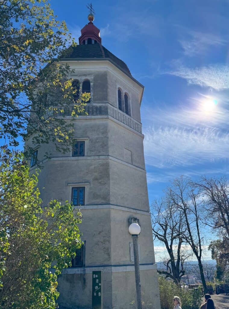 A historic tower on Schlossberg hill in Graz, with a red-tiled roof and pale stone walls. The structure is surrounded by trees, and the bright sunlight casts a warm glow on the scene.