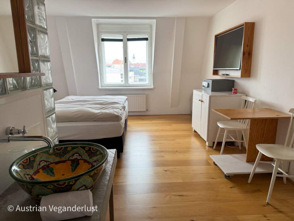 Ein helles und luftiges Hotelzimmer im Grand HOtel Wiesler in Graz Österreich mit einem großen Bett, Holzböden und minimalistischen Möbeln. Neben dem Bett steht ein buntes Keramikwaschbecken, das durch Glasbausteine abgetrennt ist. Das Zimmer hat große Fenster, die den Blick auf die Dächer der Stadt freigeben und den Raum offen und einladend wirken lassen.
