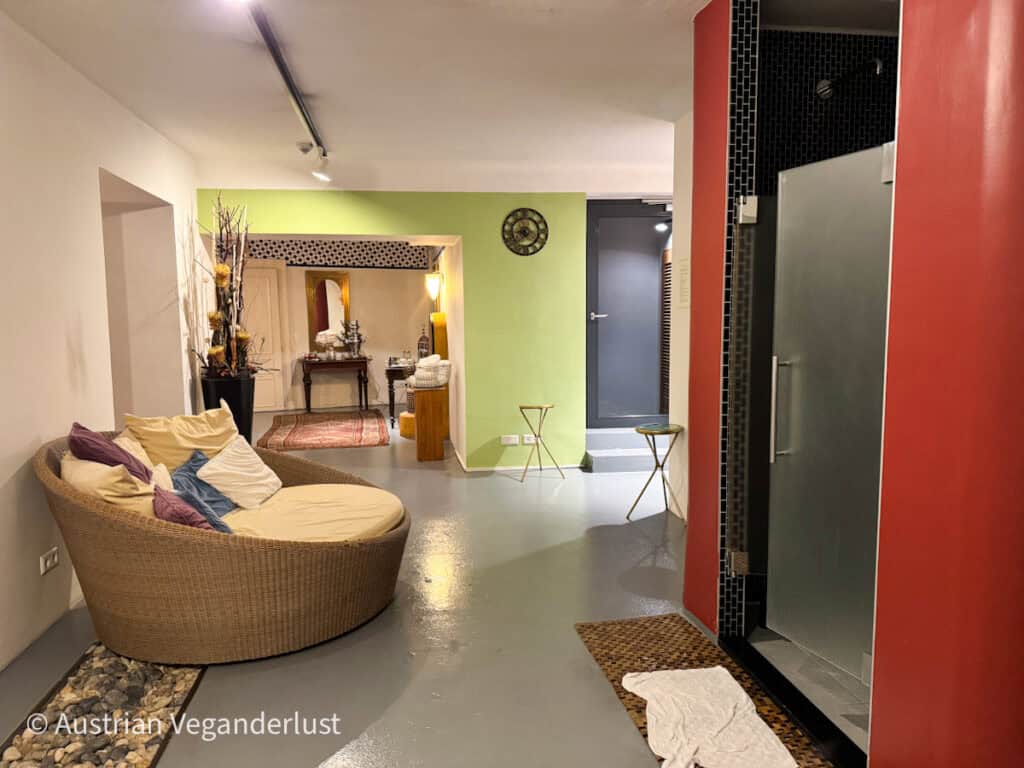 Ein entspannender Spa-Raum mit gedämpfter Beleuchtung, in dem ein Korbsessel mit Kissen im Vordergrund steht. Der Raum hat einen modernen Saunaeingang auf der rechten Seite, und die Einrichtung besteht aus Pflanzen, minimalistischen Möbeln und Erdtönen, die eine beruhigende Atmosphäre schaffen.
