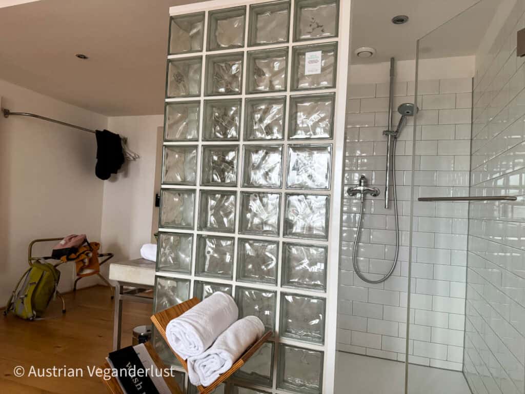 Ein minimalistisches Badezimmer mit einer Trennwand aus Glasbausteinen, die die Dusche vom Rest des Raumes trennt. Der Duschbereich ist mit weißen Subway-Fliesen und einer modernen Regendusche ausgestattet. Im Vordergrund liegen die Handtücher aufgerollt auf einem Holzständer, der das saubere und schlichte Design unterstreicht.