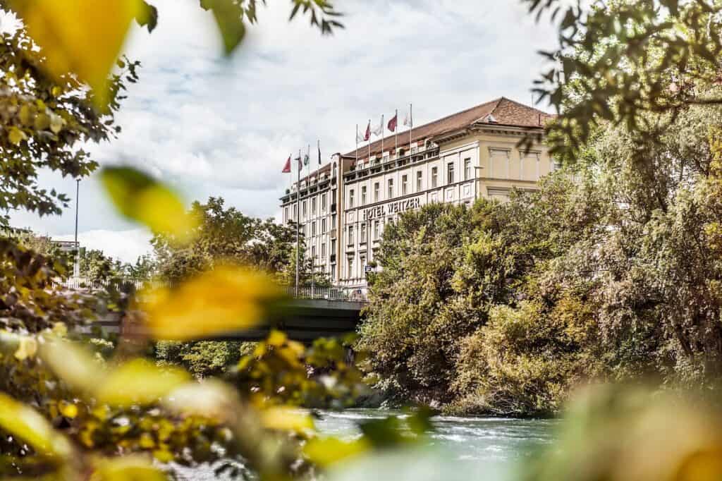 Außenansicht des Hotels Das Weitzer in Graz, Österreich. Das Gebäude ist von Bäumen umgeben und auf dem Dach wehen Fahnen. Das Hotel liegt am Flussufer, umrahmt von üppigem Grün.