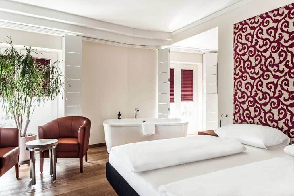 Ein helles Hotelzimmer mit einer modernen Badewanne, Ledersesseln und einer Topfpflanze. Das Zimmer hat weiße Wände und eine rot gemusterte Akzentwand, Holzböden und eine Balkontür.