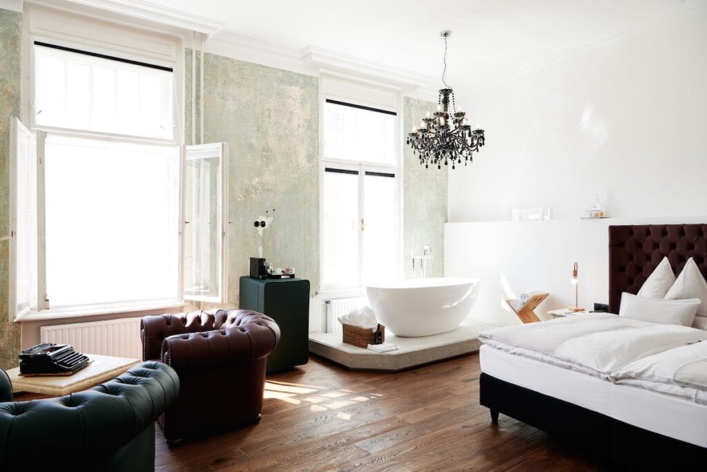 Ein stilvolles, sonnendurchflutetes Hotelzimmer mit einer freistehenden weißen Badewanne, die von zwei großen Fenstern umgeben ist. Das Zimmer ist im Vintage-Stil eingerichtet, mit dunklen Ledersesseln, einer Schreibmaschine auf einem kleinen Tisch und einem Plüschbett mit einem getufteten Kopfteil. Ein schwarzer Kristallkronleuchter verleiht dem Zimmer einen eleganten Touch, während die Holzböden und die abgenutzten Wände den raffinierten und doch rustikalen Charme des Zimmers unterstreichen.