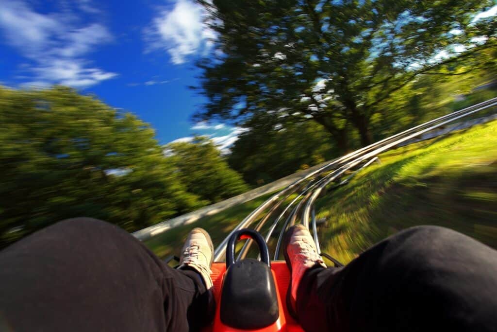 A first-person perspective from a rider on an alpine coaster, racing down a winding metal track through a lush green forest. The motion blur on the surrounding trees and blue sky creates a sense of speed and exhilaration.
