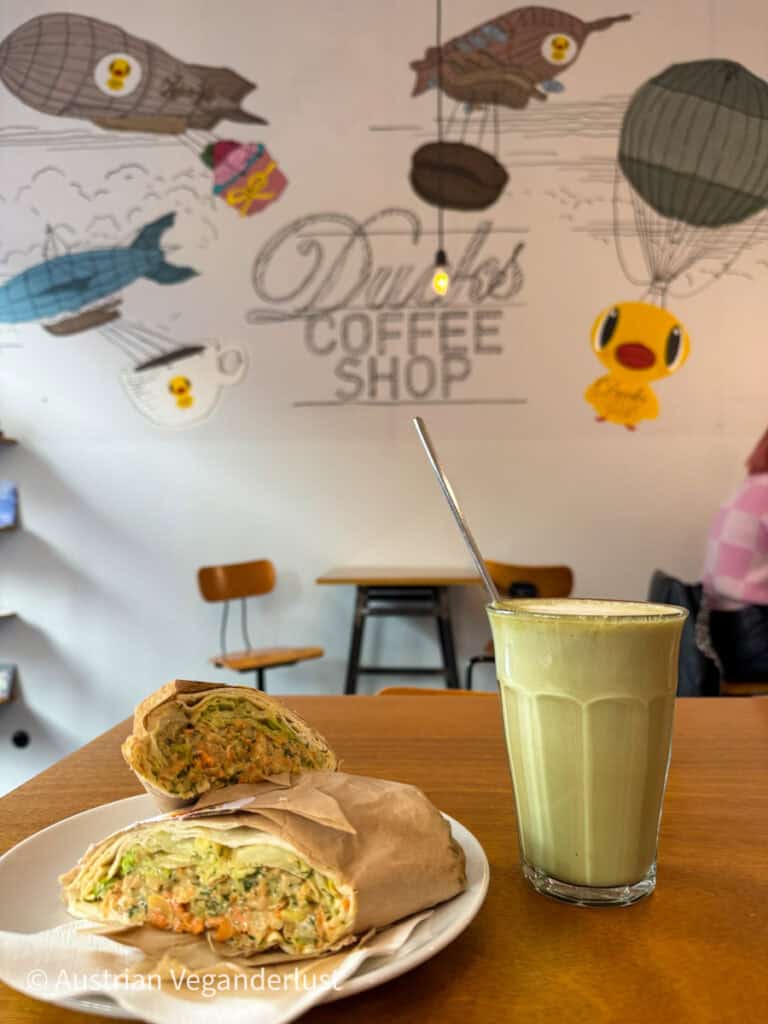 Ein Teller mit einem halbierten Gemüsewrap, auf dem die frische, bunte Füllung zu sehen ist, gepaart mit einem cremigen Matcha Latte in einem hohen Glas. Die Wand im Hintergrund ist mit verspielten Kunstwerken zum Thema Ente geschmückt, darunter schwebende Kaffeetassen und Luftschiffe.