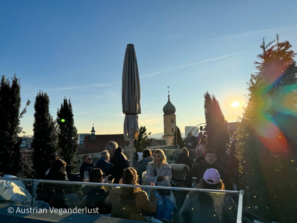 Eine Szene auf einer Dachterrasse bei Sonnenuntergang mit einer Gruppe von Menschen, die umgeben von immergrünen Topfbäumen Getränke genießen und sich unterhalten. Die Sonne wirft ein warmes Licht auf die Szene und im Hintergrund sieht man eine Kirchenkuppel und einen Kirchturm vor einem sanften blau-goldenen Himmel.
