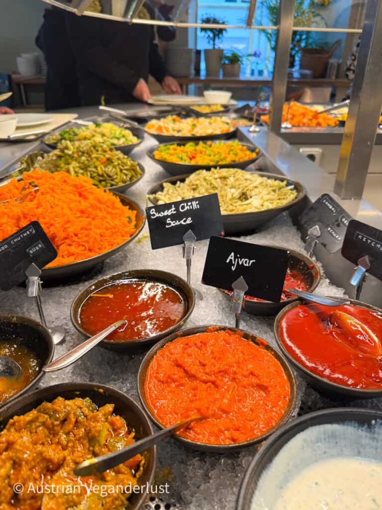 Eine lebendige Salatbar mit Schüsseln mit verschiedenen frischen Zutaten, darunter geraspelte Karotten, grüne Bohnen, gemischte Salate und Gewürze wie süße Chilisauce und Ajvar. Kleine Schilder weisen auf die Soßen hin, während im Hintergrund ein legeres Ambiente mit Tellern und Gästen zu sehen ist.