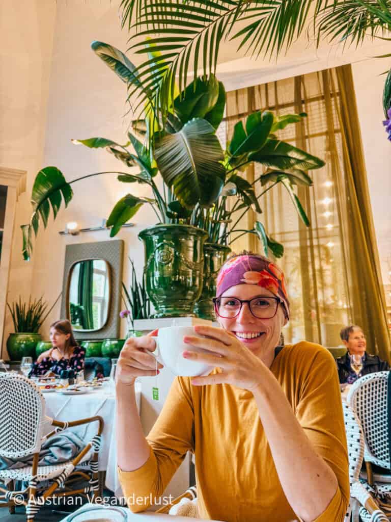Eine Frau mit Brille und buntem Kopftuch lächelt, während sie eine große weiße Teetasse in einem schicken, mit Pflanzen gefüllten Café hält. Sie trägt ein senffarbenes Oberteil und sitzt vor einem üppigen grünen Pflanzenarrangement, das die entspannte, tropische Atmosphäre des Raums noch verstärkt. Andere Gäste sitzen im Hintergrund und genießen ihre Mahlzeiten, was die gemütliche Atmosphäre noch verstärkt.