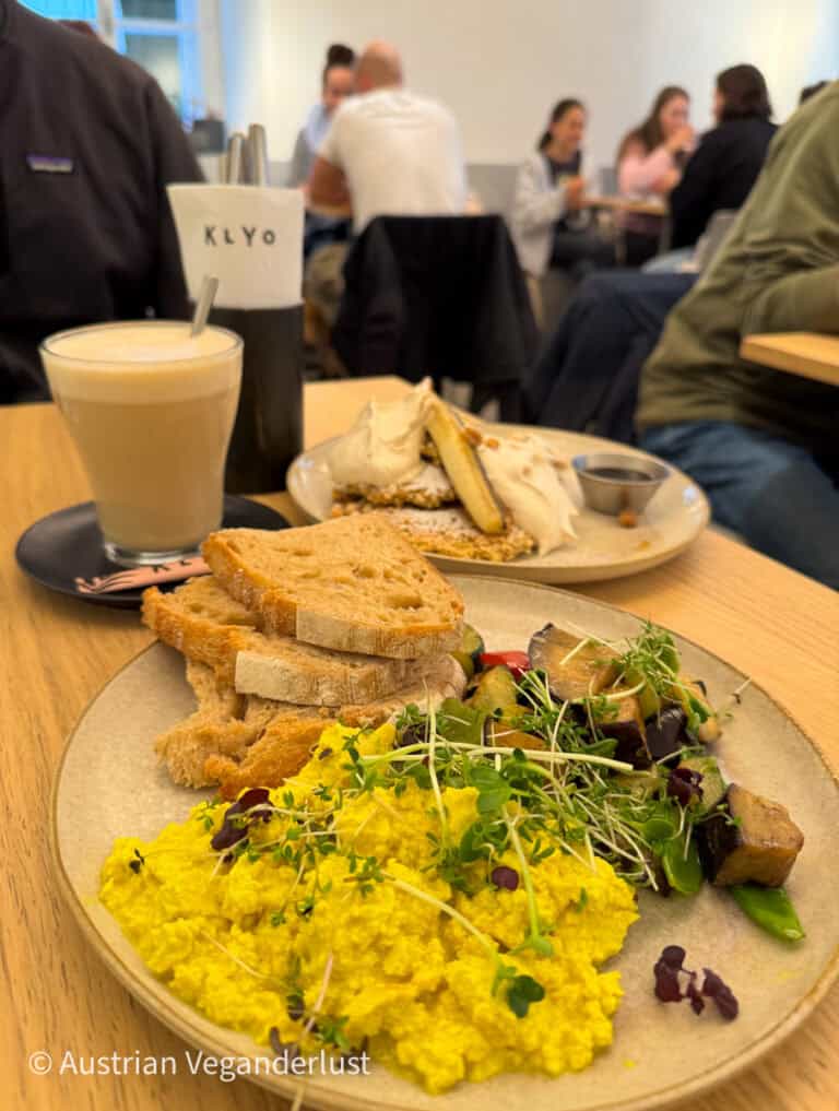 A plate of vibrant scrambled tofu with fresh sprouts, roasted vegetables, and slices of rustic bread, accompanied by a latte. Diners are visible in the café's relaxed and bustling atmosphere, enhancing the friendly ambiance.