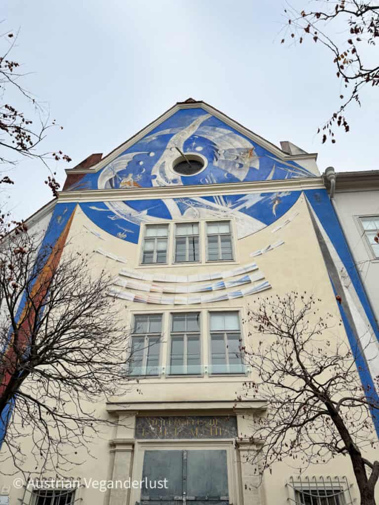 Eine lebendige, großflächige Sonnenuhr schmückt die Fassade eines Gebäudes mit verschlungenen blauen und weißen Mustern, die himmlische und astronomische Elemente darstellen. Im Mittelpunkt der Gestaltung steht ein rundes Fenster in der Nähe der Spitze. Die gemalten Linien und Symbole strahlen nach außen und enthalten künstlerische Darstellungen von Sternen und Planeten. Laublose Baumzweige umrahmen die Szene und verleihen diesem architektonischen Kunstwerk einen saisonalen Touch.