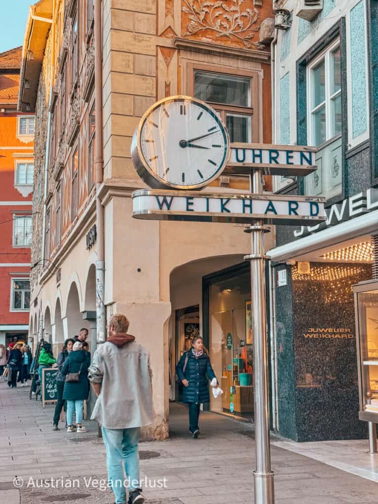 The Weikhard clock in Graz, Austria, prominently displayed on a polished metal post with "Uhren Weikhard" signage beneath its clean, modern clock face. The scene captures a bustling street with shoppers walking past historic buildings featuring intricate architectural details and arches, set against the warm light of late afternoon.