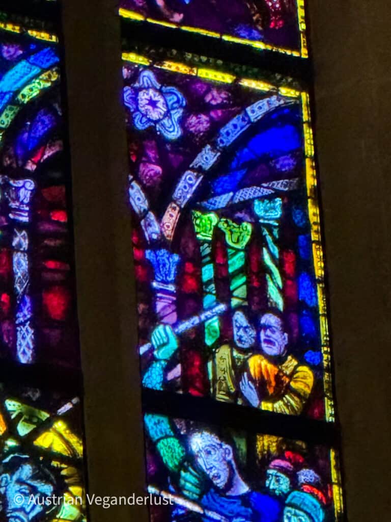 A close-up of a stained glass window in a church, showcasing detailed depictions of figures and vivid colors, including blues, purples, and yellows. The artwork features expressions and gestures that convey a dramatic biblical narrative.
