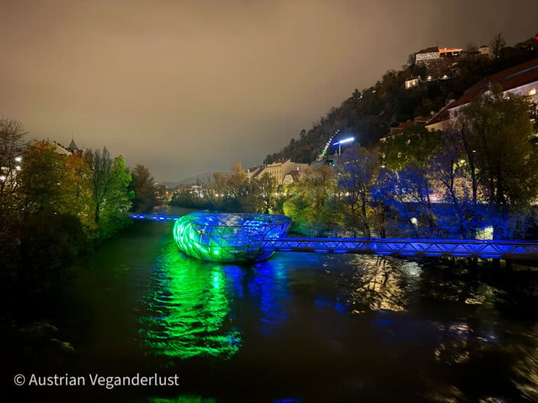 Die beleuchtete Murinsel, eine schwimmende Stahlkonstruktion in Form einer Muschel auf der Mur in Graz, Österreich, leuchtet mit grünen und blauen Lichtern und wirft bunte Reflexionen auf das Wasser. Die Bäume in der Umgebung und die nahe gelegenen Gebäude sind sanft beleuchtet und unterstreichen die ruhige nächtliche Atmosphäre.
