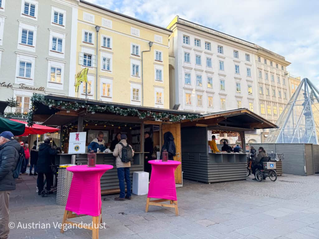 Dieses Bild zeigt einen festlichen Weihnachtsmarktstand auf dem Alten Markt in Salzburg, umgeben von historischen, in Pastelltönen gestrichenen Gebäuden. Leuchtend rosafarbene Tischdecken verleihen dem Ganzen einen modernen Touch, während sich die Besucherinnen und Besucher versammeln, um Speisen und Getränke zu genießen. Eine gläserne Pyramide im Hintergrund verleiht der Szenerie einen architektonischen Reiz.