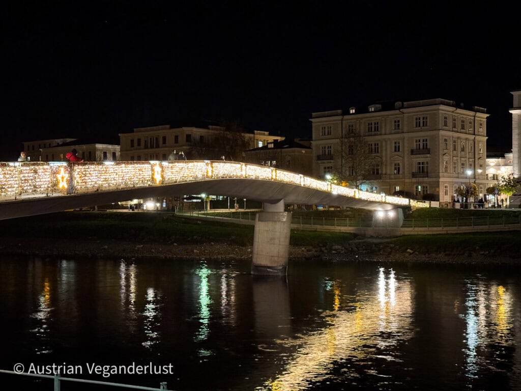 A brightly illuminated pedestrian bridge in Salzburg, decorated with Christmas lights and star motifs, reflecting beautifully on the Salzach River below. Historic buildings along the riverbank complete the serene winter night scene.