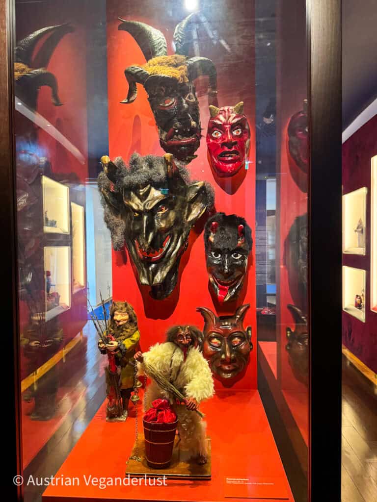 A collection of traditional Krampus masks and figurines displayed in a red-lit case at the Salzburg Christmas Museum. The intricate details of the masks capture the folkloric character's eerie and dramatic essence.
