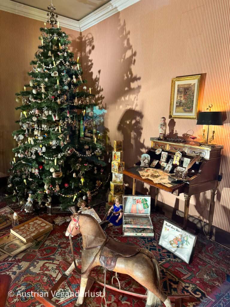A cozy, vintage Christmas scene from the Salzburg Christmas Museum, featuring a beautifully decorated tree with ornaments and candles, surrounded by antique toys like a wooden rocking horse and dolls. The room evokes the warmth and charm of old-fashioned holiday celebrations.