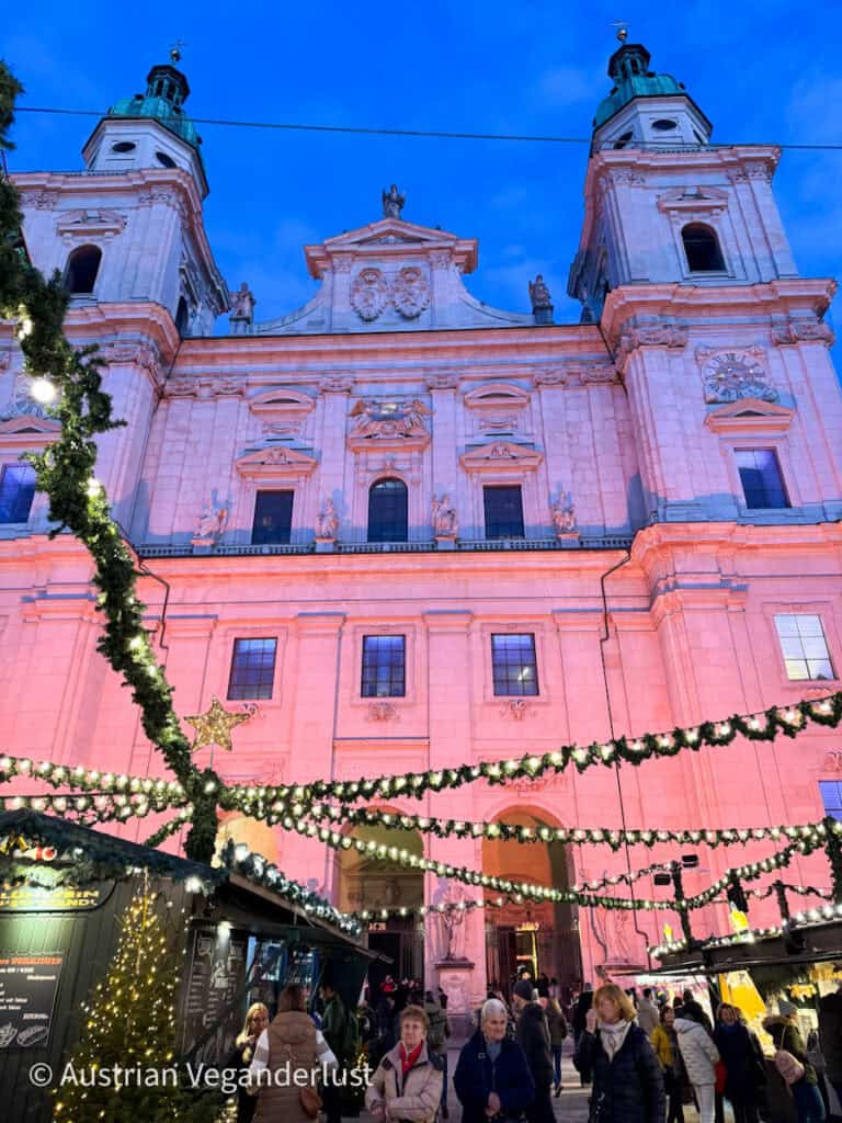 Der wunderschön rosa beleuchtete Salzburger Dom überragt den belebten Weihnachtsmarkt am Domplatz. Über den Marktständen hängen Lichterketten, die eine gemütliche, festliche Atmosphäre schaffen, während die Besucher die weihnachtlichen Angebote entdecken.