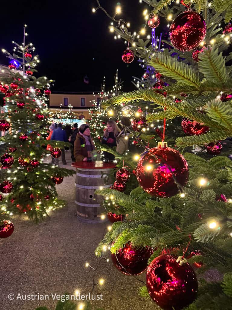 A close-up view of Christmas trees adorned with glowing lights and shiny red ornaments at the Hellbrunn Christmas Market in Salzburg. Visitors are gathered around a barrel-style table in the background, enjoying the festive atmosphere.