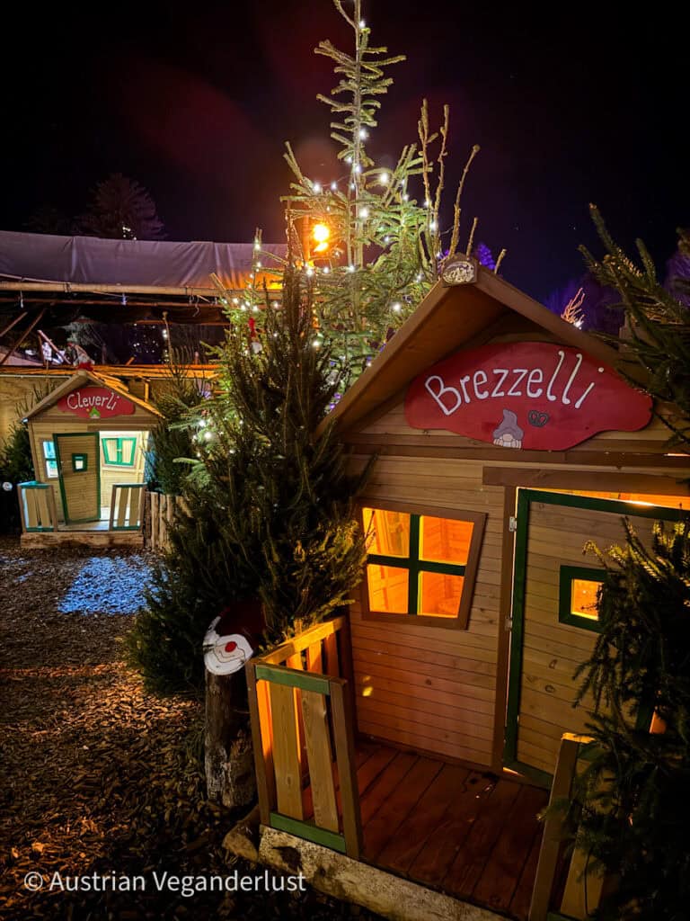 Auf dem Hellbrunner Christkindlmarkt in Salzburg stehen bezaubernde hölzerne Spielhäuser, die mit "Cleverli" und "Brezzeli" bezeichnet werden, umgeben von beleuchteten Weihnachtsbäumen. Die gemütliche Szene strahlt weihnachtliche Wärme und Laune aus.