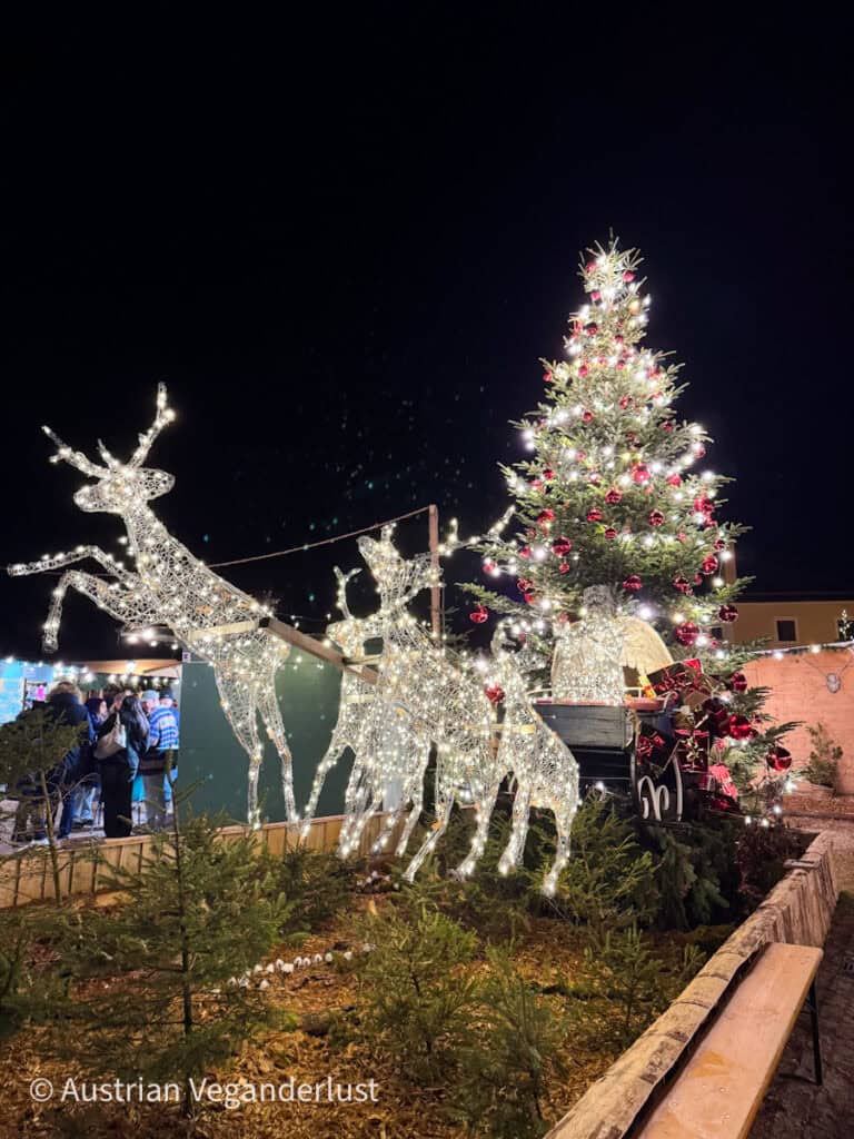 Beleuchtete Rentierfiguren ziehen einen dekorativen Schlitten neben einem festlich beleuchteten und mit roten Ornamenten geschmückten Weihnachtsbaum auf dem Hellbrunner Weihnachtsmarkt. Die Besucher im Hintergrund tragen zur lebhaften Weihnachtsatmosphäre bei.