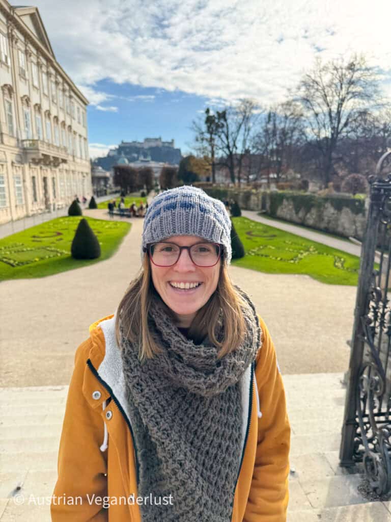 A cheerful visitor wearing a knit hat and scarf poses in front of the Mirabell Gardens in Salzburg. The beautifully manicured garden paths and hedges lead to a stunning view of Hohensalzburg Fortress perched on a hill in the background under a partly cloudy sky.