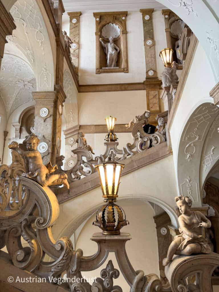 A grand staircase in Mirabell Palace, Salzburg, with ornate marble railings, cherub statues, and classical sculptures in alcoves. Warmly lit lamps add to the luxurious and artistic ambiance of the baroque architecture.