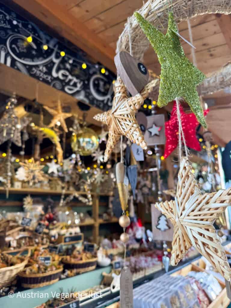 Eine Nahaufnahme von handgefertigtem Weihnachtsschmuck, darunter Weidengeflecht und glitzernde Sterne, die an einem Marktstand am Mirabellplatz hängen. Warme Lichter und eine Vielzahl von Weihnachtsartikeln schaffen eine gemütliche, festliche Atmosphäre.