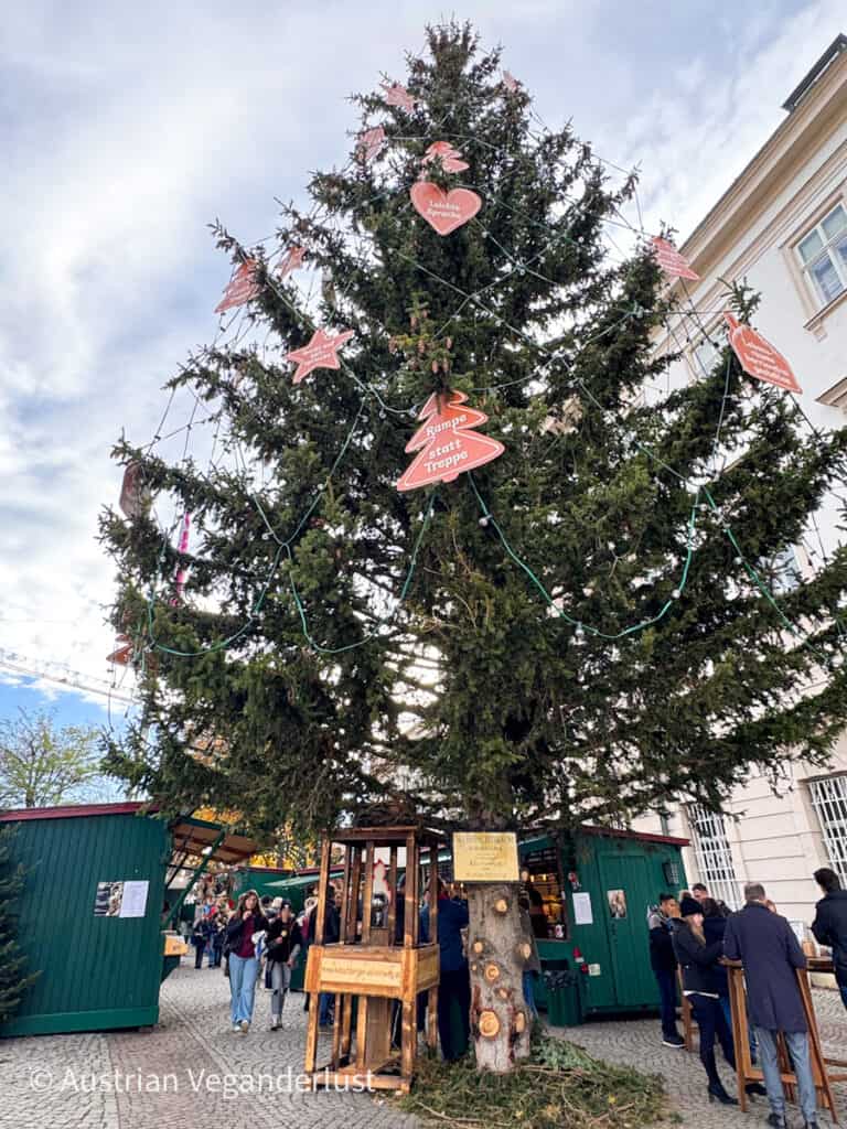 Ein hoher Weihnachtsbaum, geschmückt mit Ornamenten und Schildern mit Botschaften, steht auf dem Weihnachtsmarkt am Mirabellplatz. Der Baum ist von grünen Holzbuden umgeben, die weihnachtliche Waren und Leckereien anbieten, und die Besucher erkunden die Gegend.