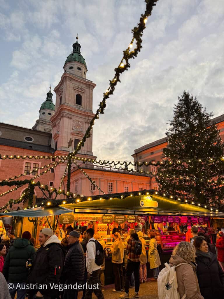 A busy market stall at Residenzplatz in Salzburg, selling a variety of festive treats and gifts, under strings of illuminated garlands. The Salzburg Cathedral looms in the background, softly lit as the sky transitions to evening.