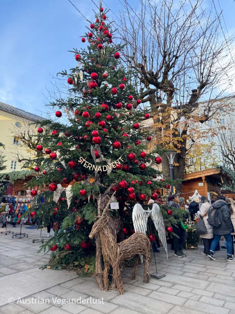 Ein großer, mit leuchtenden Lichtern und glänzenden roten Ornamenten geschmückter Weihnachtsbaum steht an prominenter Stelle auf dem Stern-Adventsmarkt in Salzburg. Unter dem Baum sorgen eine rustikale Rentier-Skulptur aus Stroh und Engelsflügel für einen festlichen, natürlichen Touch, während die Besucher den Markt durchstöbern.