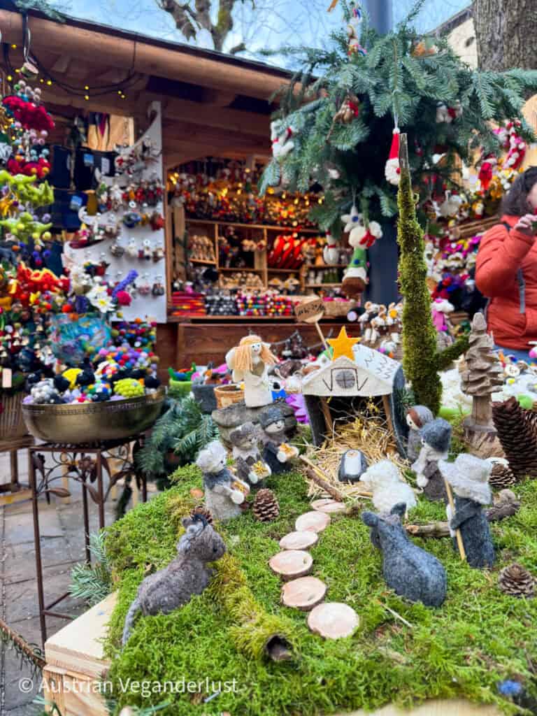 Eine detailgetreue Krippe mit handgefertigten Tier- und Menschenfiguren auf einem moosbedeckten Ständer auf dem Stern-Adventsmarkt. Im Hintergrund sorgt ein Stand mit farbenfrohem Weihnachtsschmuck und Dekorationen für eine fröhliche Weihnachtsatmosphäre.