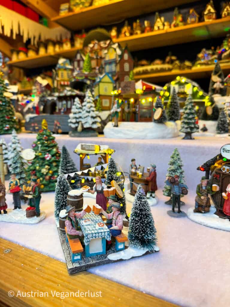 Ein lebhaft dekoriertes Miniatur-Weihnachtsdorf mit winzigen schneebedeckten Häusern, Bäumen und festlichen Figuren, die weihnachtlichen Aktivitäten nachgehen, wie zum Beispiel in einem Biergarten essen. Blinkende Lichter verleihen der Szene Wärme und Charme.