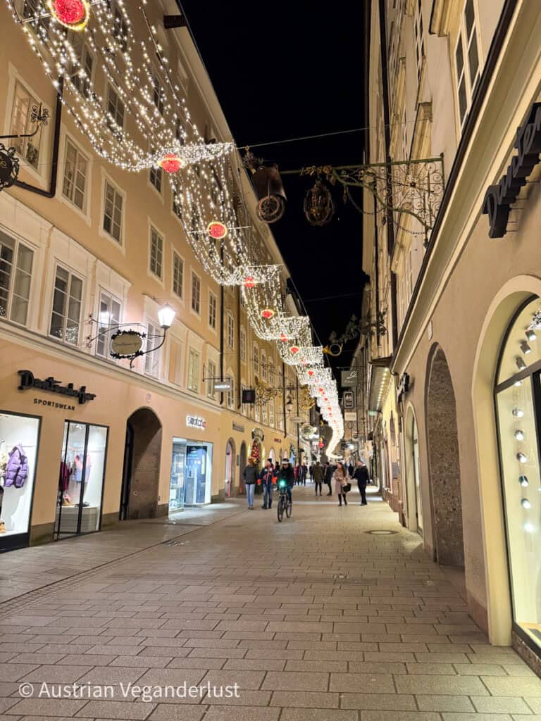 Getreidegasse in Salzburg, adorned with sparkling Christmas lights and red decorations hanging above the historic street. Shoppers and visitors stroll past charming storefronts, enjoying the festive atmosphere.
