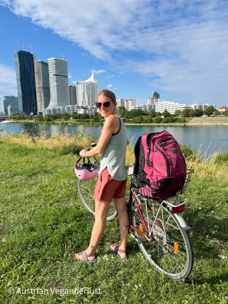Tina steht mit ihrem Fahrrad auf einer Wiese am Flussufer in Wien. Sie trägt einen rosafarbenen Helm und hat einen großen Rucksack im hinteren Korb des Fahrrads. Moderne Hochhäuser aus der Skyline der Wiener Donau City ragen unter einem teilweise bewölkten Himmel über die Donau.