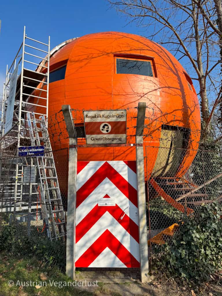 Ein leuchtend orangefarbenes, kugelförmiges Gebäude, das als "Republik Kugelmugel" bekannt ist, liegt hinter einem Stacheldrahtzaun mit einem rot-weiß gestreiften Tor; Schilder weisen es als Grenzübergang der Mikronation in Wien aus.