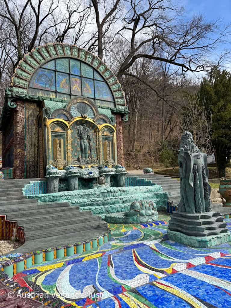 Das kunstvolle Äußere von Otto Wagners Villa in Wien mit dem großen Bogeneingang, den kunstvollen Bronzeskulpturen und dem stufenförmigen Brunnen, umrahmt von üppigen Bäumen und lebhaften Mosaikfliesen in wirbelnden Mustern.