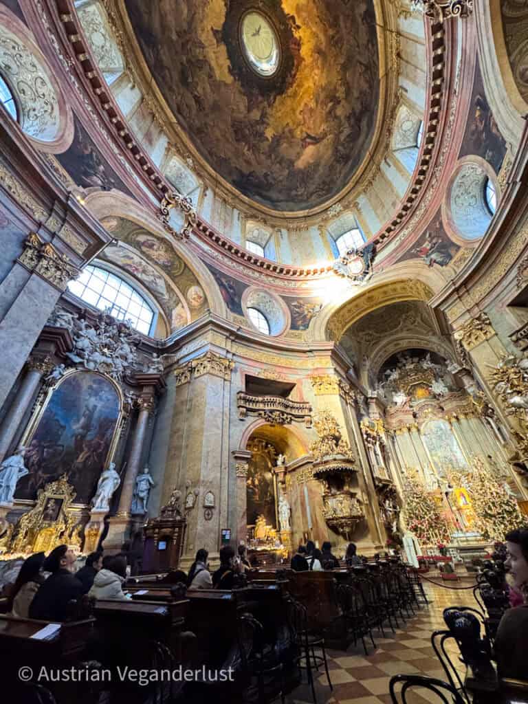 Das opulente Innere der Peterskirche in Wien zeigt ihre großartige barocke Architektur. Die hoch aufragende Kuppel ist mit kunstvollen Fresken geschmückt, während vergoldete Skulpturen und verzierte Altäre den reich verzierten Raum füllen. Die Besucher sitzen in dunklen Holzbänken und bewundern die goldene Kanzel und den beleuchteten, festlich geschmückten Altar.
